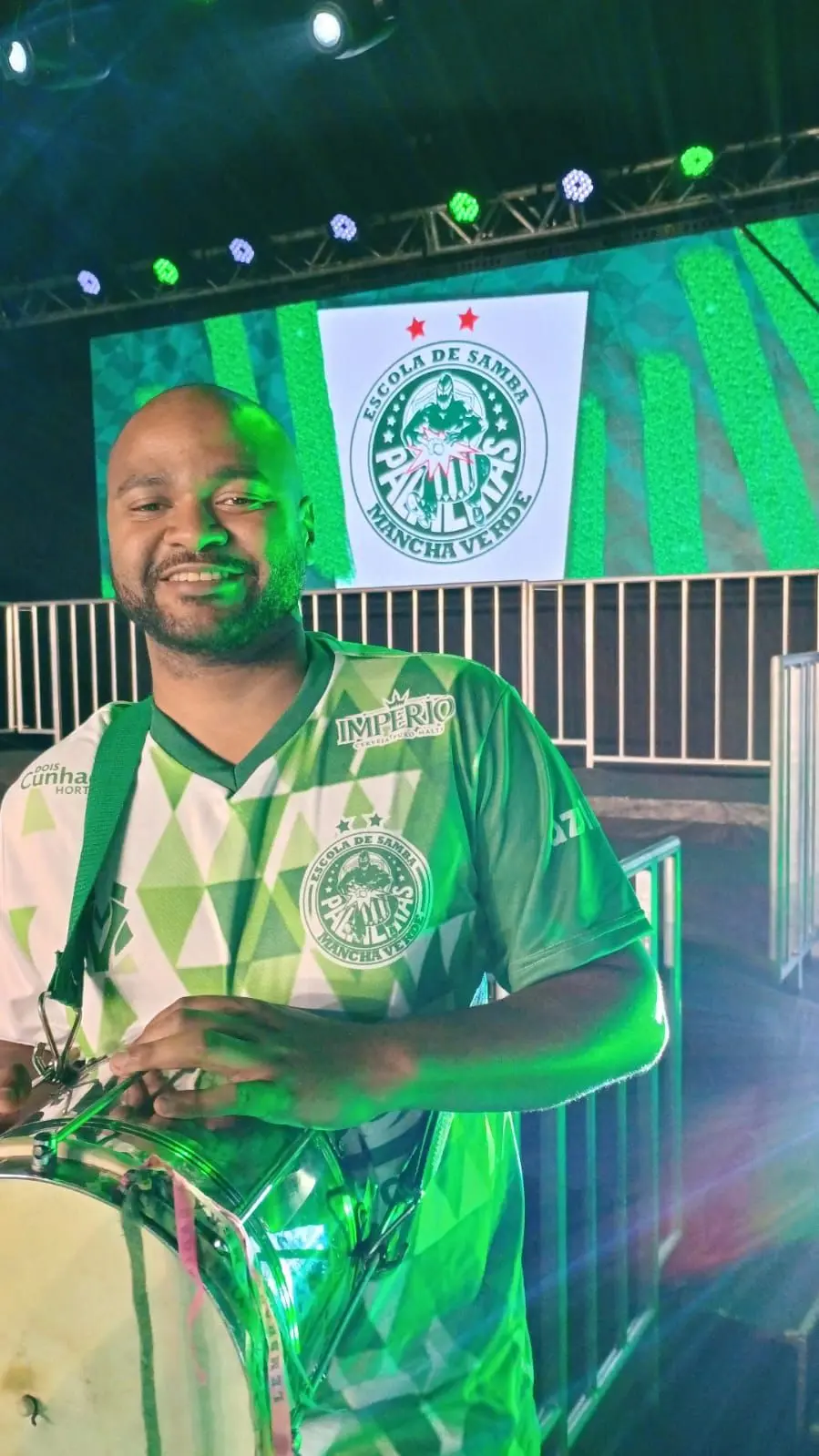 Deyvidson Machado Andrade tocando cuíca em palco, com o símbolo da Mancha Verde ao fundo